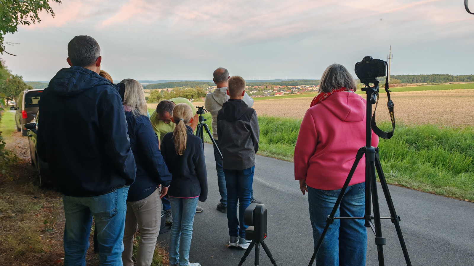 Eine Gruppe von Menschen steht auf einer Straße mit Kameras und Stativen, die auf eine weite Landschaft gerichtet sind.