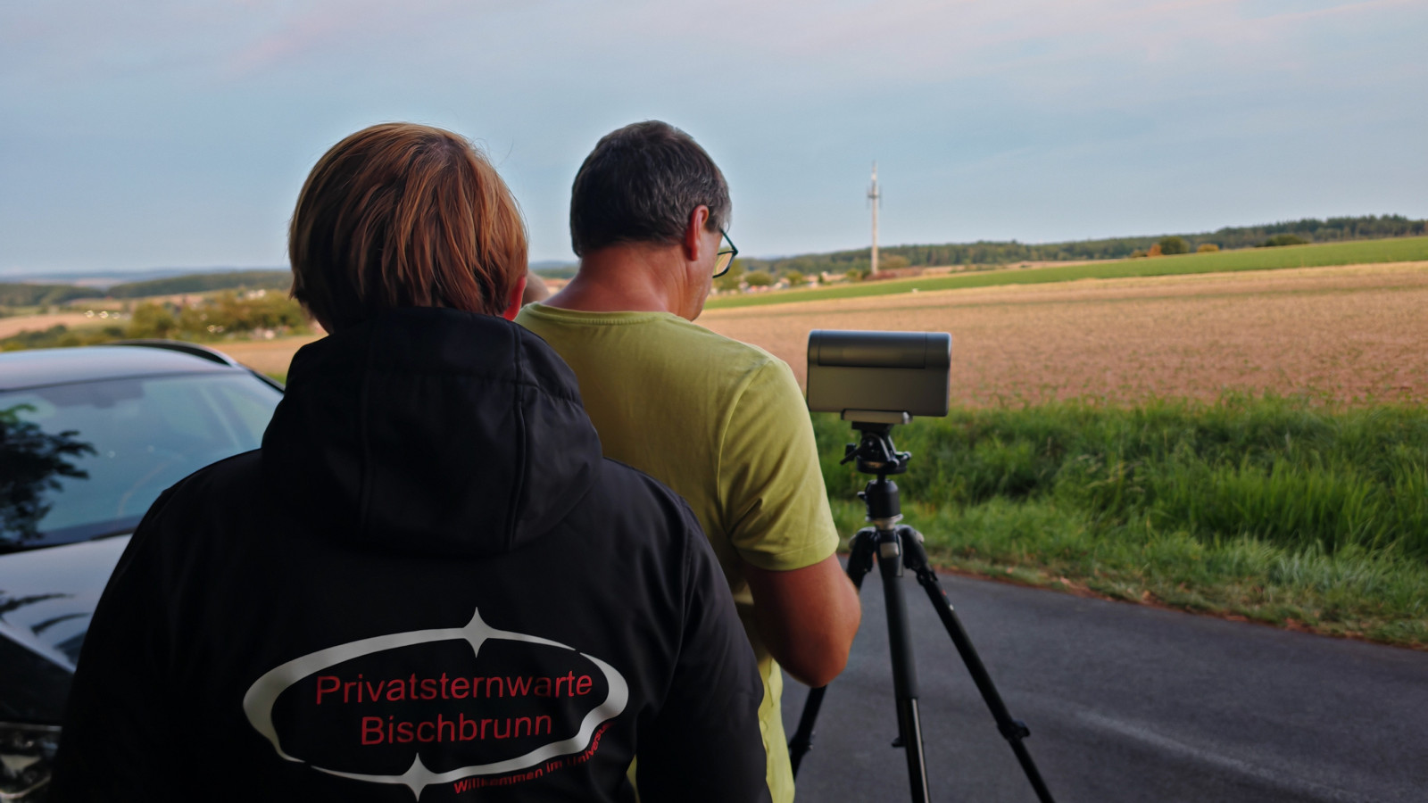 Zwei Personen schauen durch ein Teleskop am Straßenrand bei Abenddämmerung.